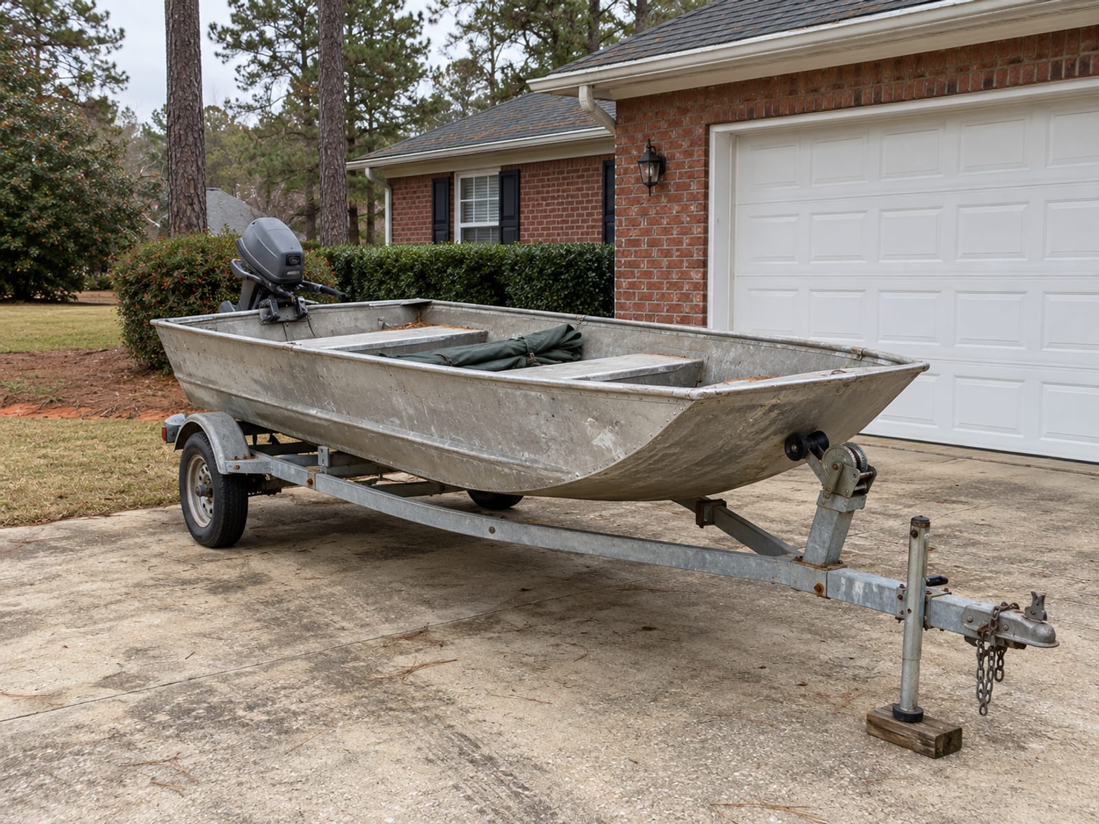 A small aluminum fishing boat on a trailer parked beside a suburban garage
