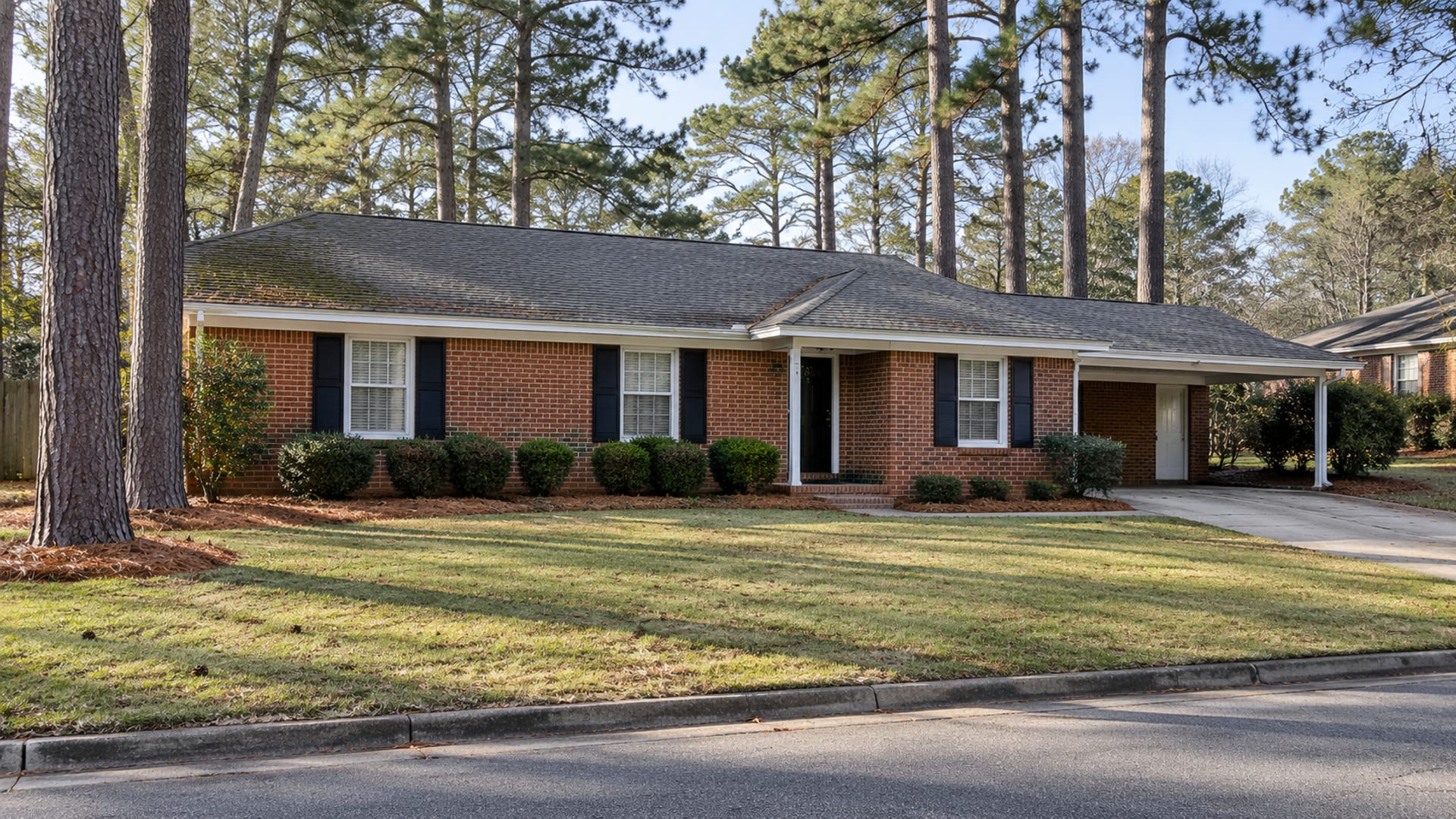 A brick ranch house with a shingle roof under tall pines on a quiet Alabama street