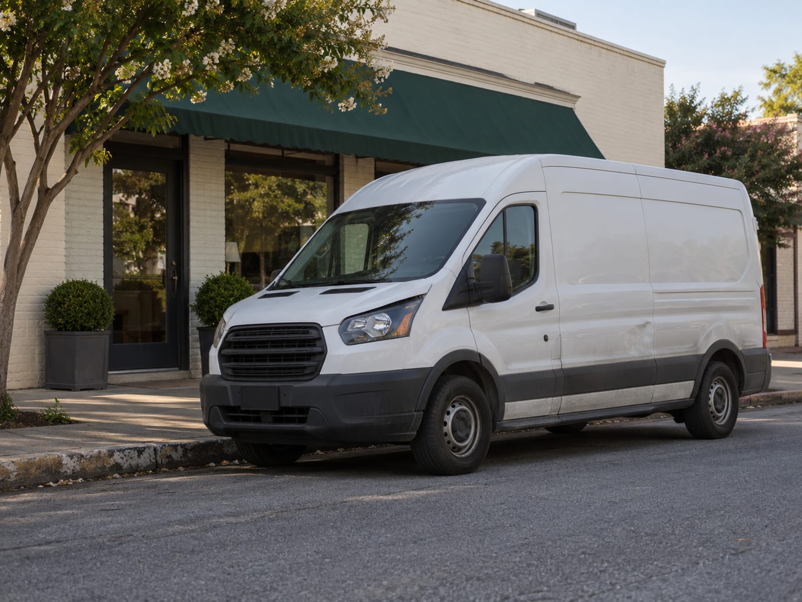 A plain white cargo van parked at a curb beside a small storefront in the morning