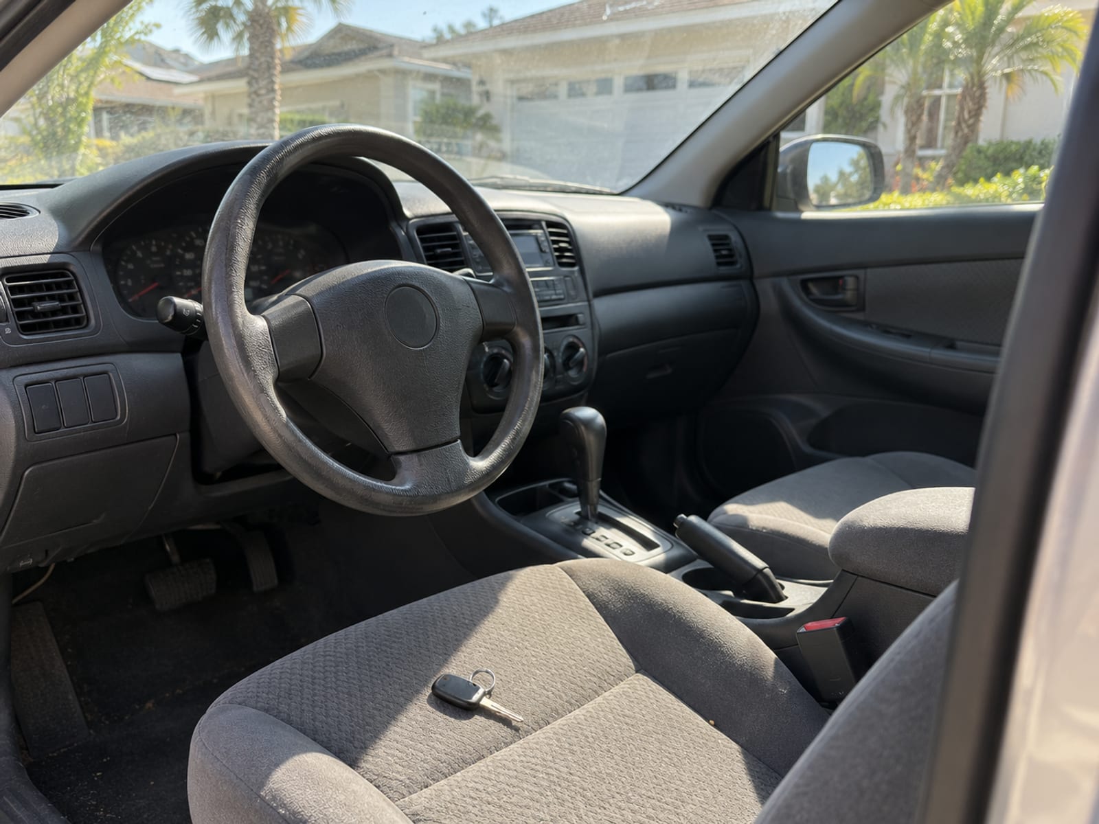 The steering wheel and empty driver's seat of a parked car in a sunny Florida driveway, keys resting on the seat and the dashboard out of focus