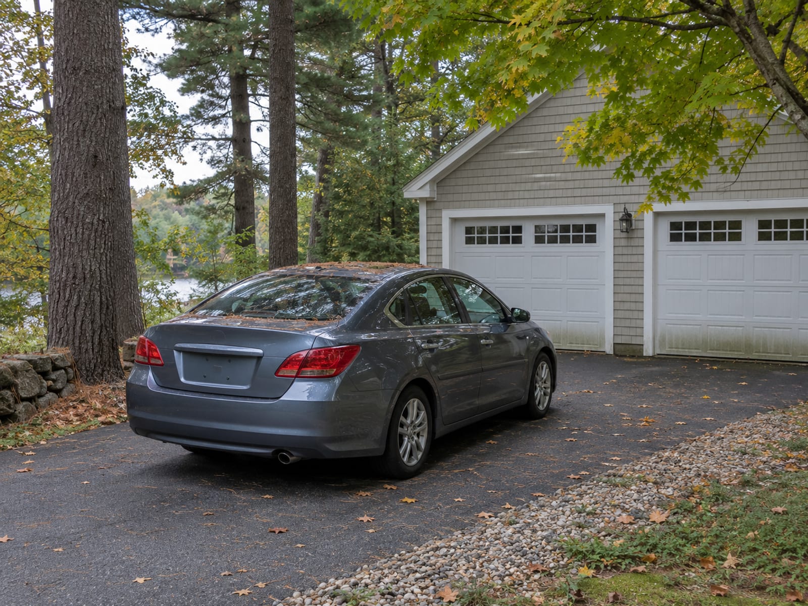 A sedan parked in a gravel driveway beside a barn-red garage under maple trees