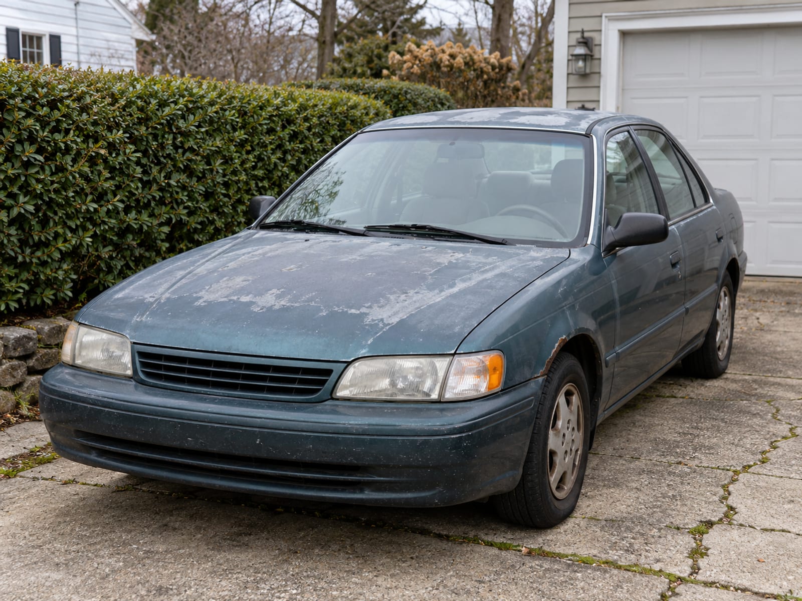 An older sedan with faded paint parked in a driveway beside a hedge