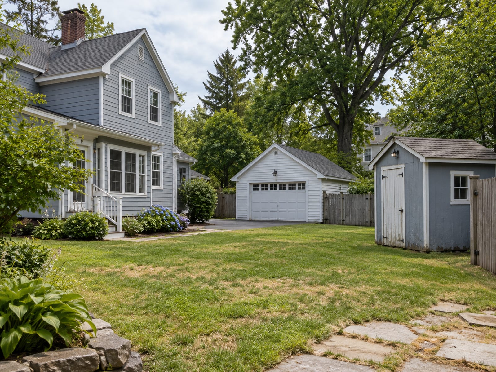 A New England home with a detached garage, a small garden shed and a wooden fence in the yard