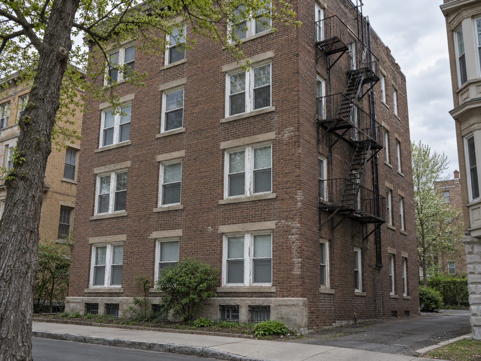 A brick 1920s apartment building with painted wooden window trim and a fire escape on a city side street