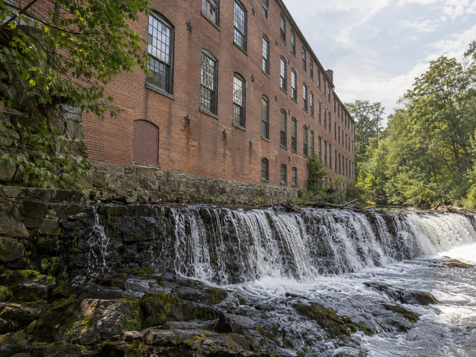 A long brick former mill building with rows of tall windows beside a narrow river and a stone dam