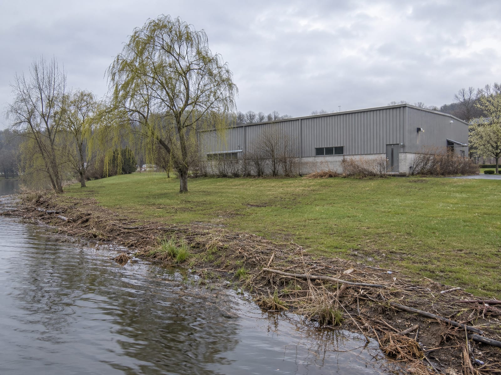 A low commercial building and a parking lot beside a slow river with a grassy floodplain bank on an overcast day