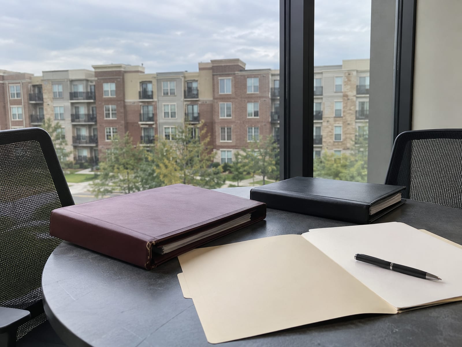 A conference table with closed binders, a pen and a blank policy folder beside a window overlooking apartment buildings
