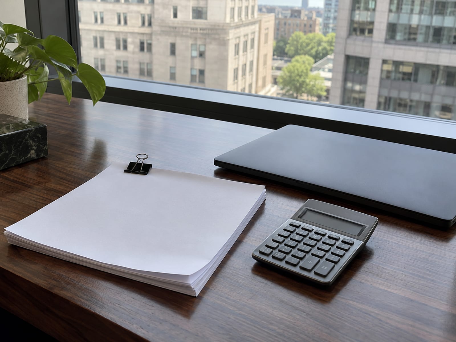 A desk with a closed laptop, a calculator and a stack of blank invoice sheets beside a window overlooking city office buildings