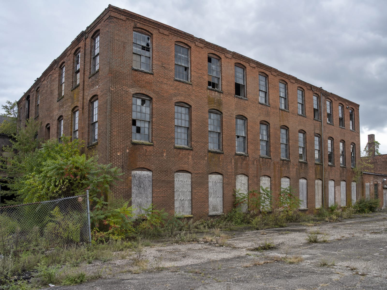 A vacant two-story brick warehouse with boarded lower windows beside an empty paved lot under an overcast sky