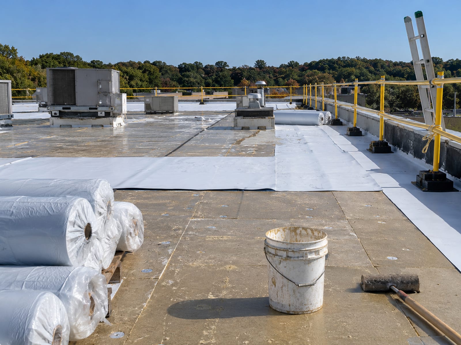 A flat commercial roof with new membrane rolls, a ladder and safety rails under a clear sky