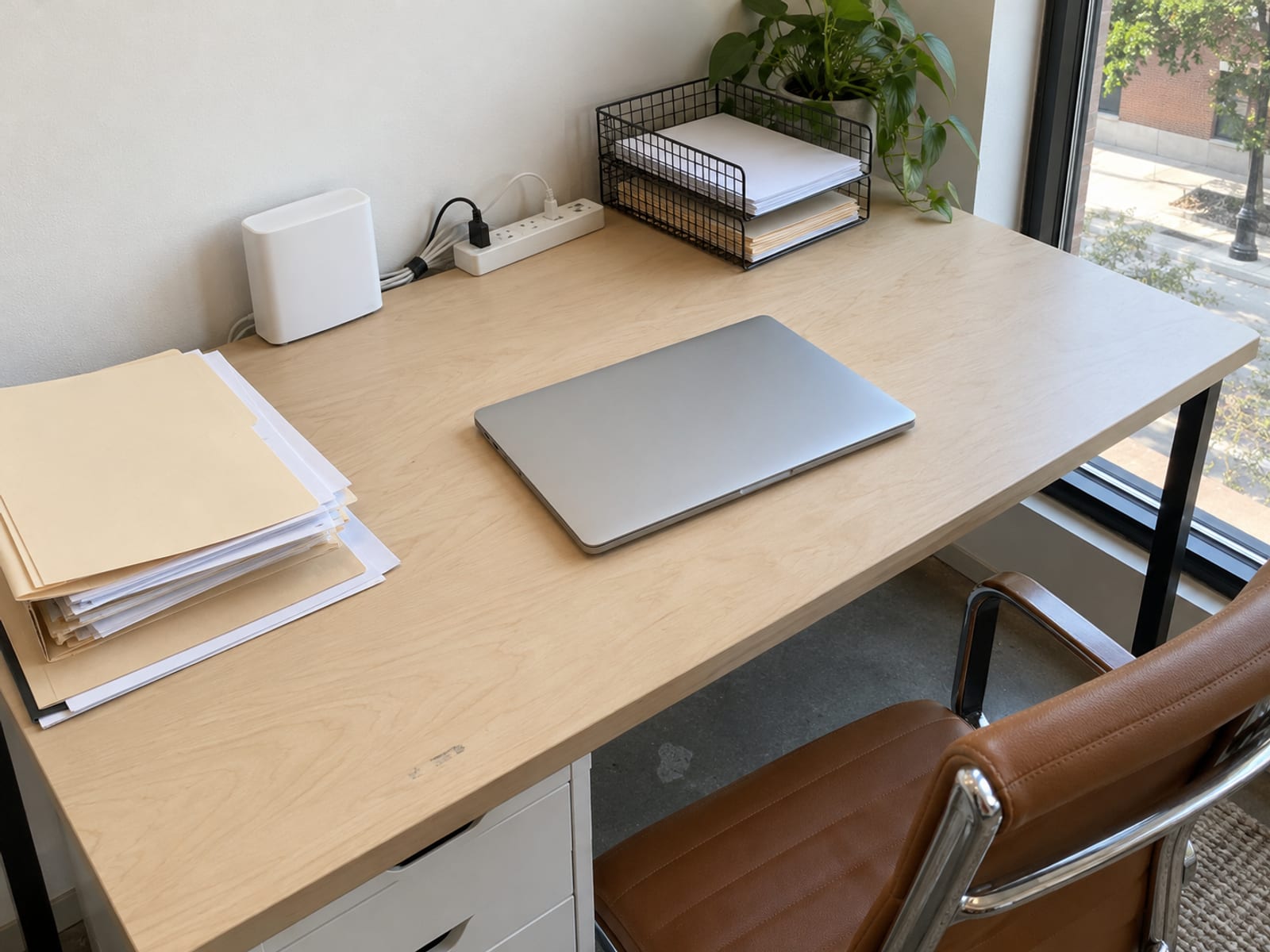Un escritorio pequeño de oficina con una laptop cerrada, un router y una pila de expedientes en papel bajo luz suave de día
