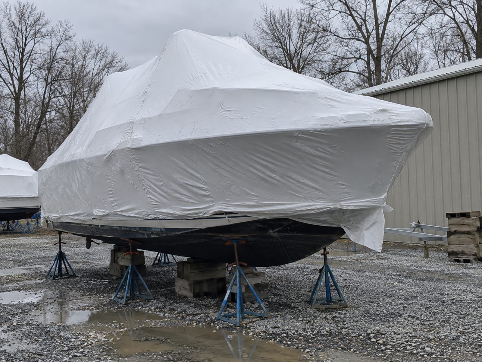 A shrink-wrapped boat on stands in a boatyard during winter with bare trees behind it