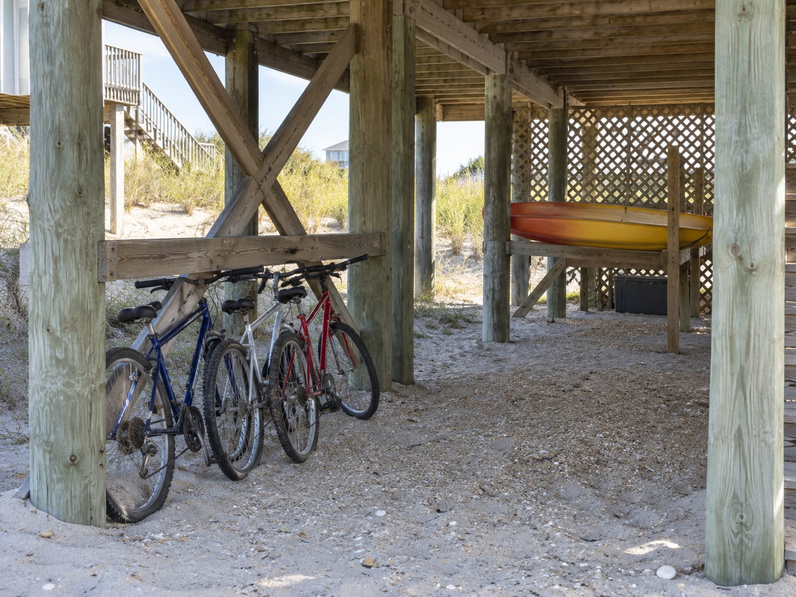 The open ground-level area beneath a raised beach house on pilings, with a few stored bikes and a kayak