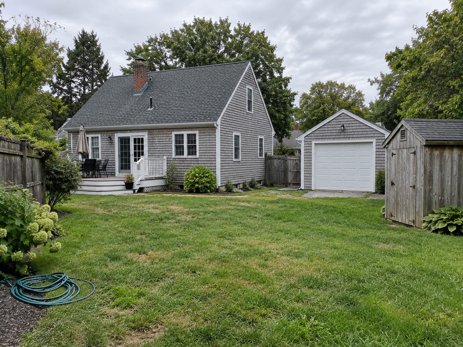 A New England style house with a detached garage, a small garden shed and a wooden fence in the yard