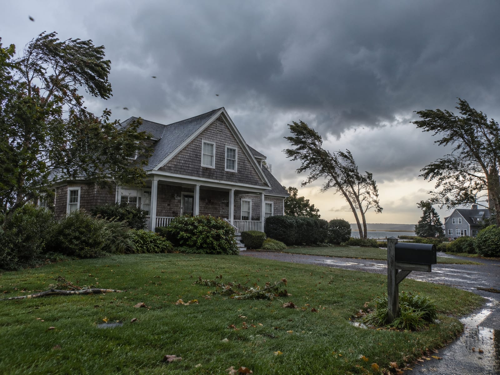 A shingled coastal home under gathering storm clouds with bending trees and a wet lawn