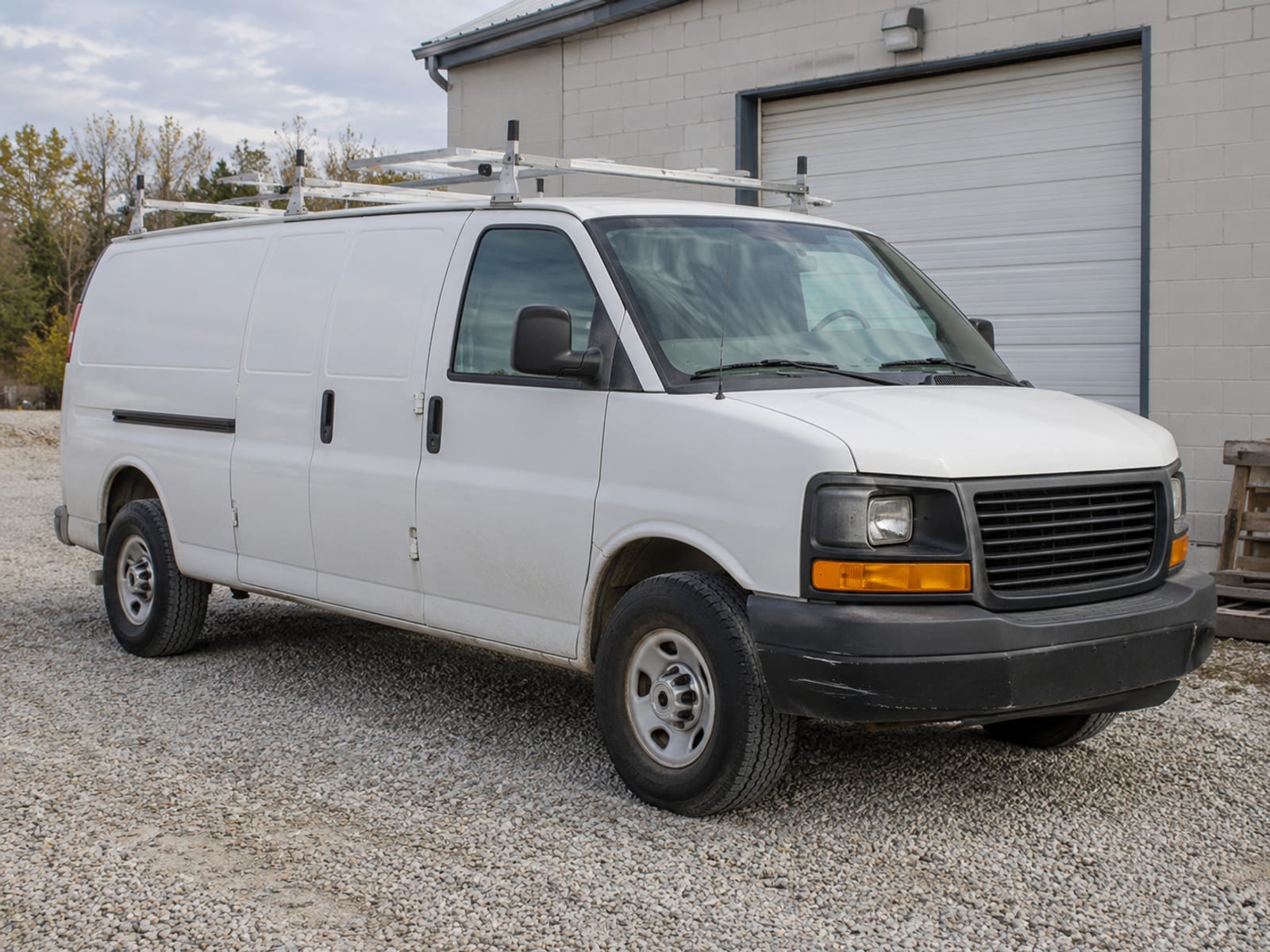A plain white work van with a ladder rack parked outside a small workshop