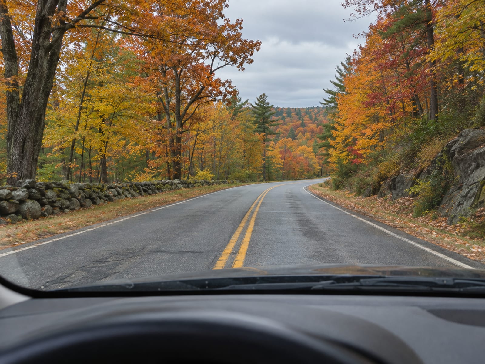 Una carretera vacía de dos carriles que se curva entre árboles de otoño, vista a través del parabrisas de un auto