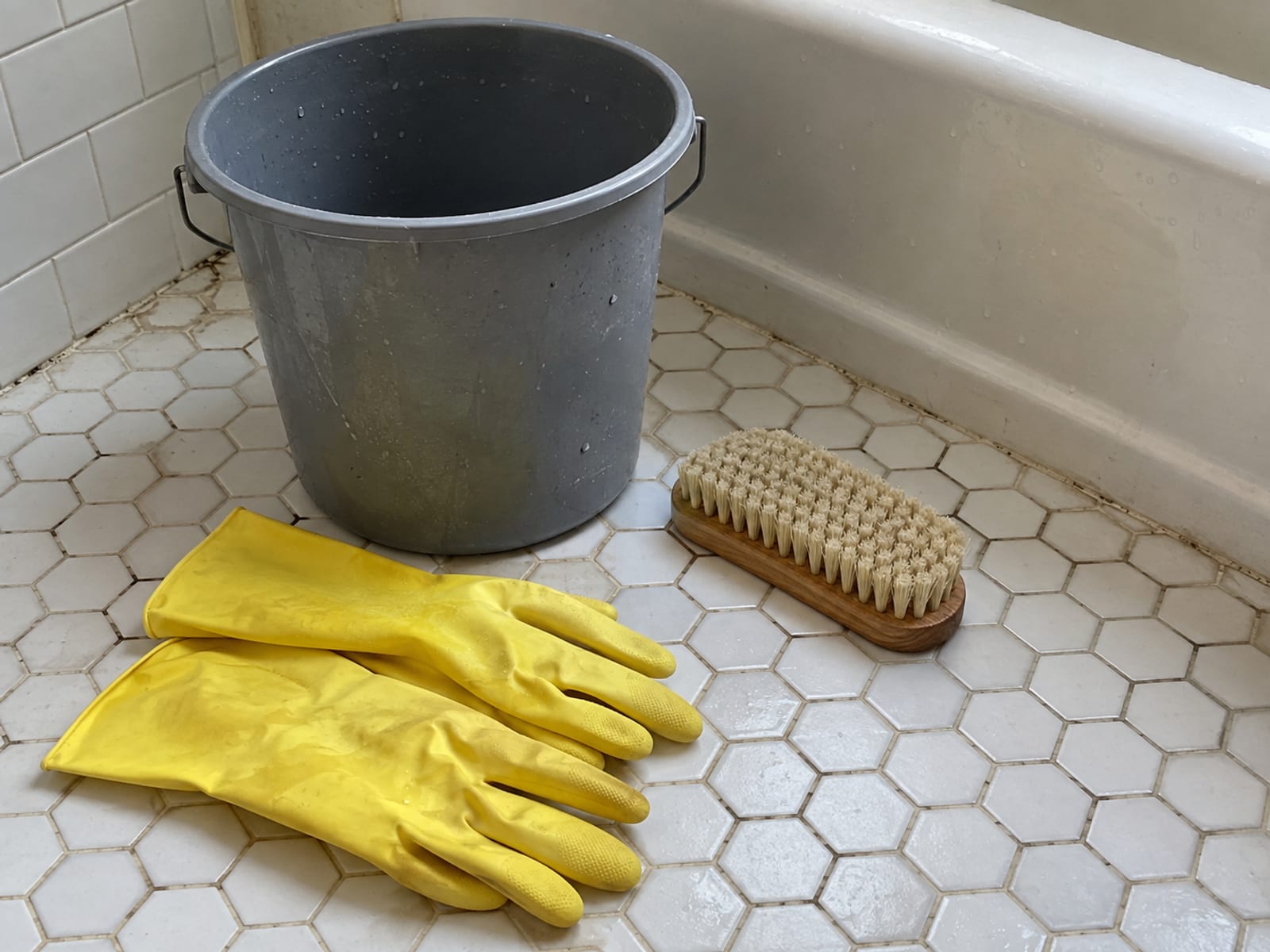 A bucket, a scrub brush and a pair of rubber gloves on a tiled bathroom floor