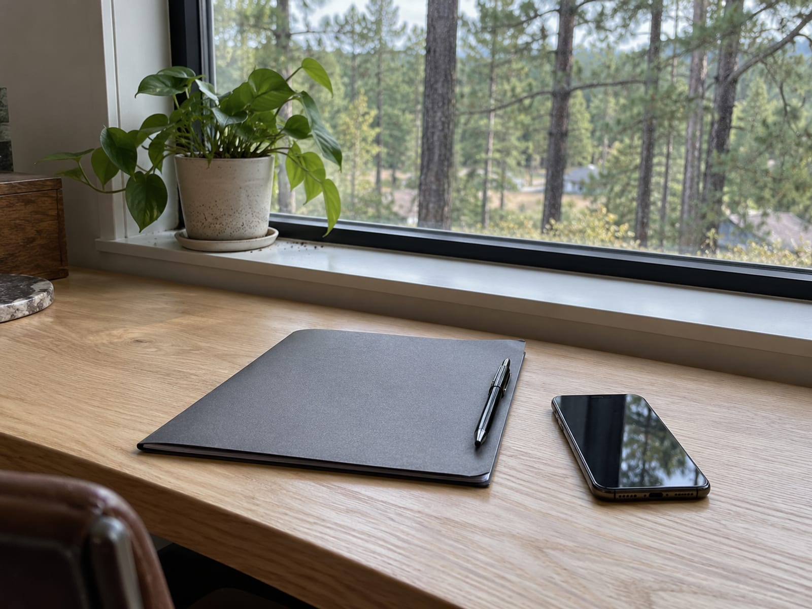 A desk with a closed folder, a pen and a phone beside a window with soft afternoon light