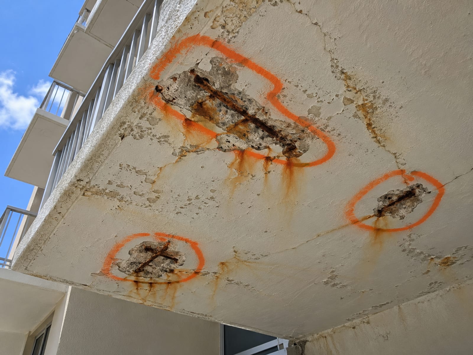 A concrete balcony underside with exposed rebar marked for repair on a coastal building