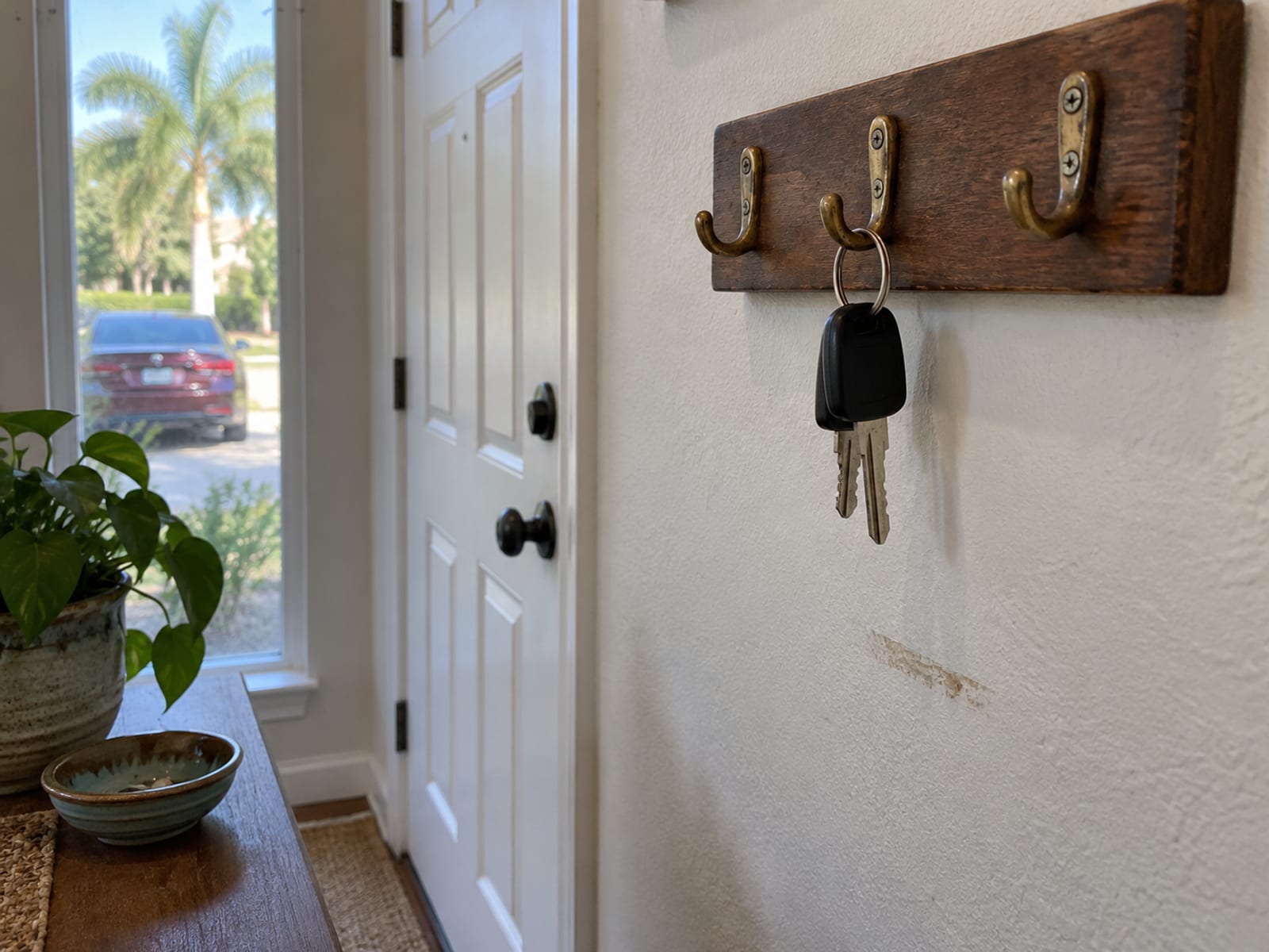 A plain set of car keys hanging from a wall hook beside a front door, a parked car blurred through the window