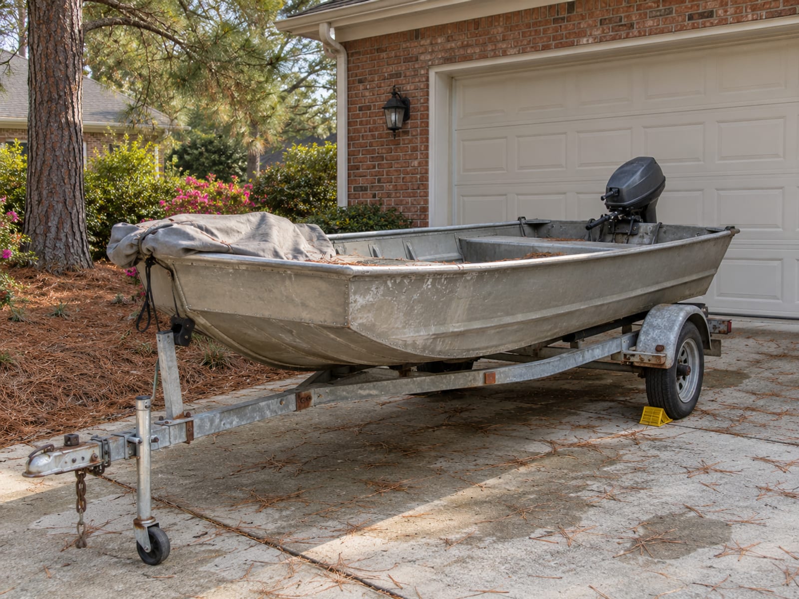 A small aluminum fishing boat on a trailer parked beside a suburban garage