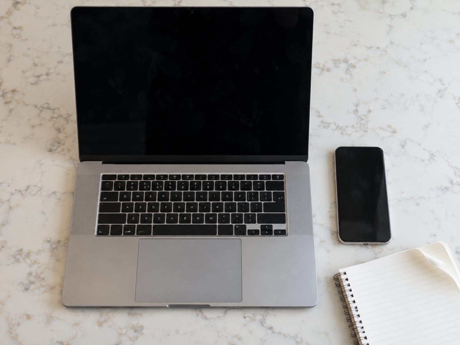 A laptop and a phone side by side on a desk beside a blank notepad