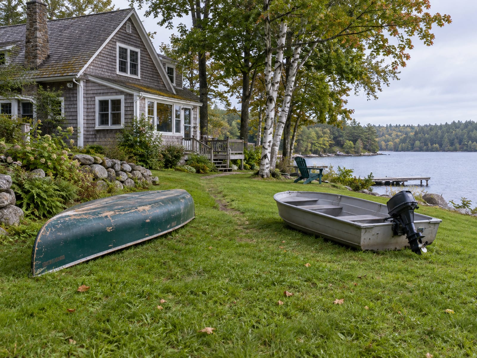 A canoe and a small outboard boat resting on the lawn beside a shingled house near a lake
