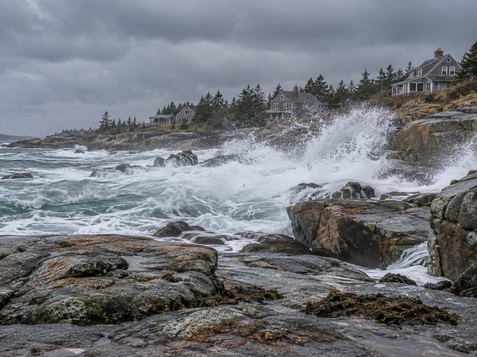 Waves washing over a rocky shoreline toward shingled cottages under a gray storm sky