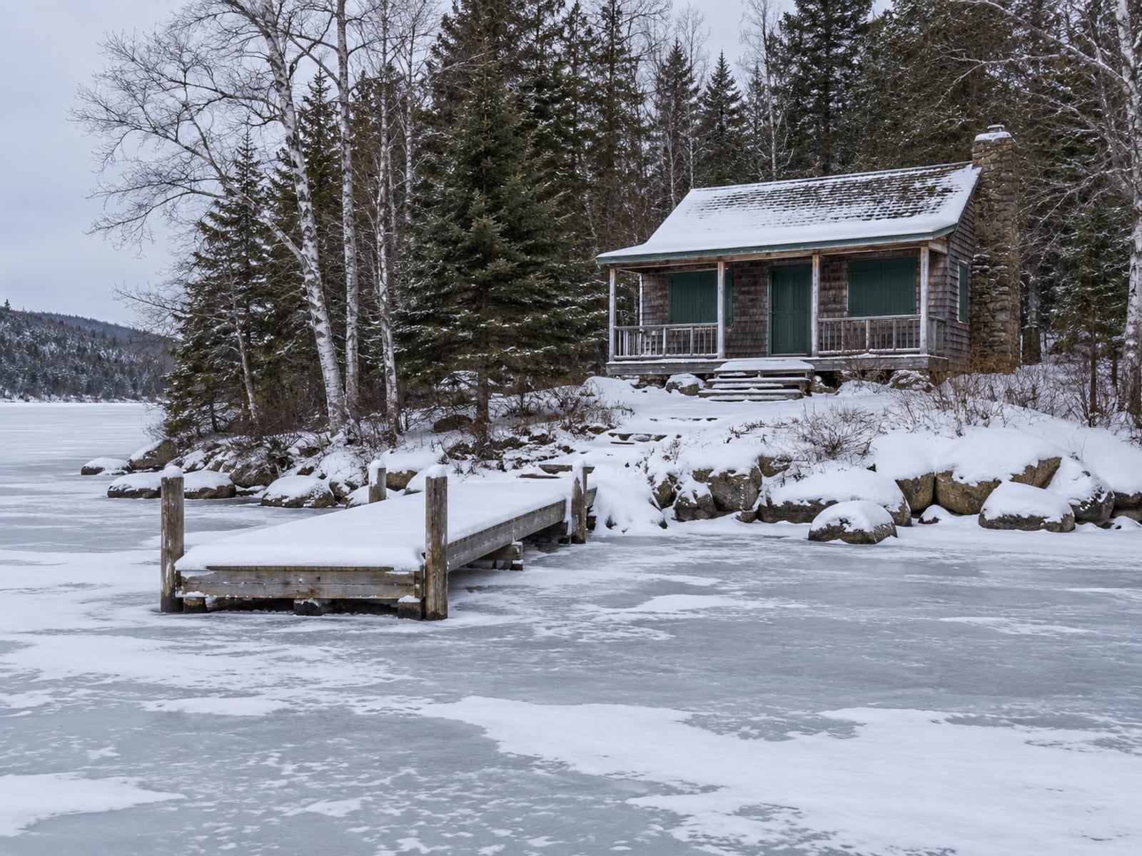 A small wooden camp on a lakeshore with its windows shuttered and snow on the dock