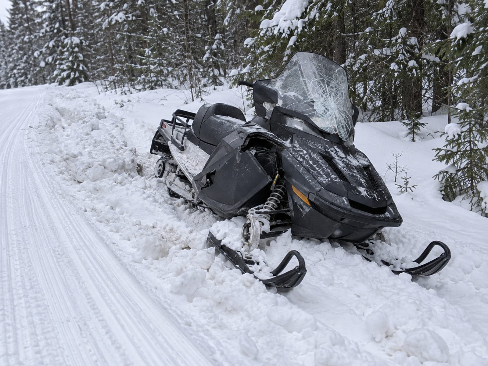 A snowmobile stopped at the side of a wooded trail with a bent ski resting in the snow