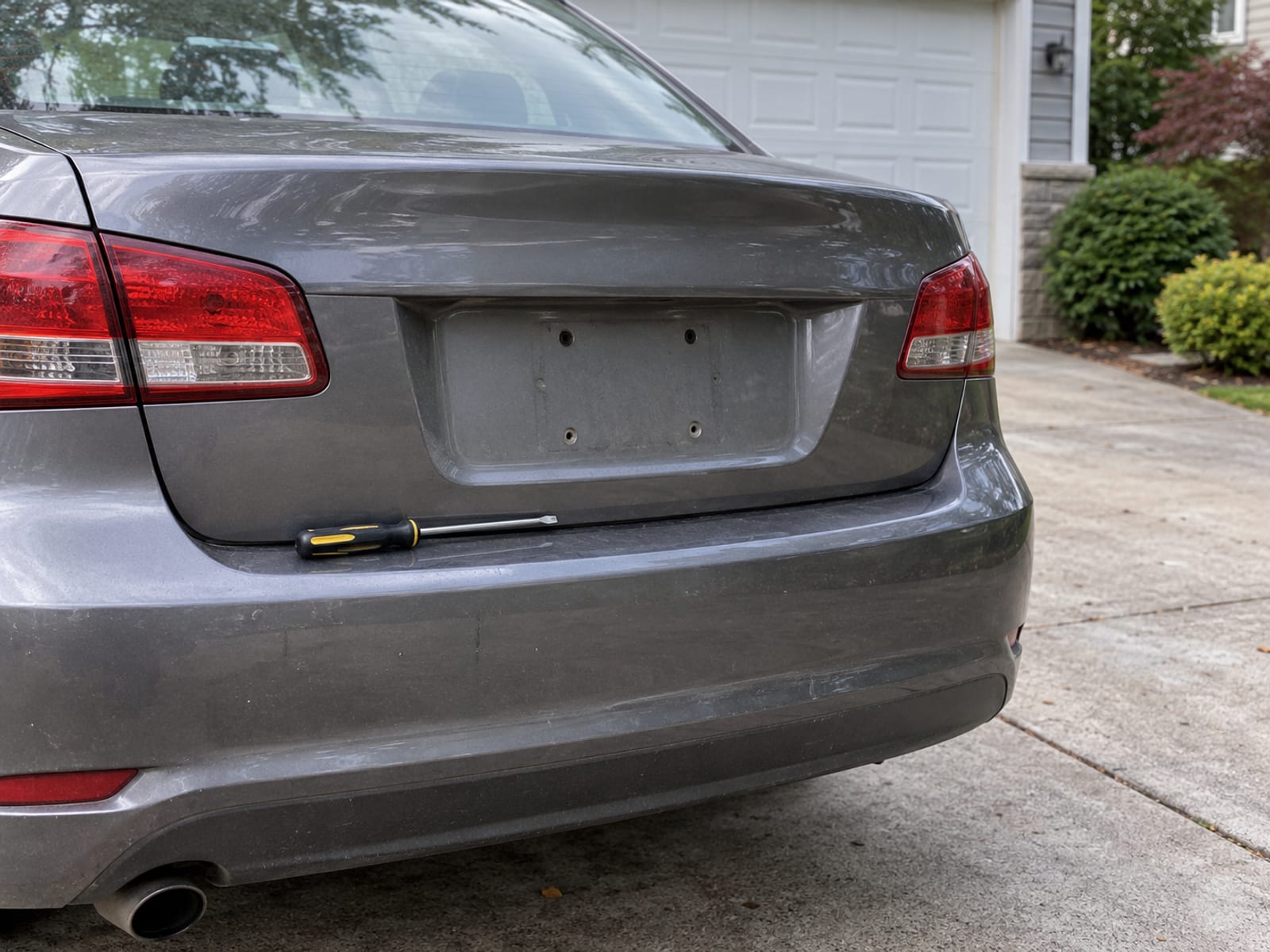 The rear of a sedan in a driveway with its license plate removed and a screwdriver resting on the bumper