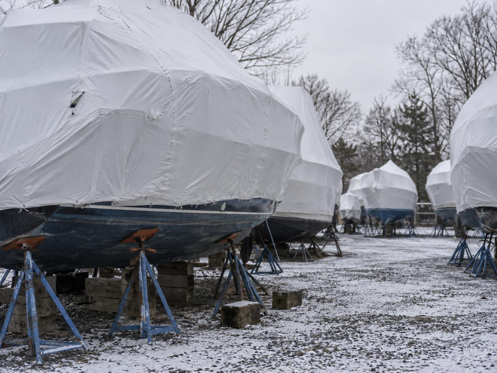 Several shrink-wrapped boats stored on stands in a boatyard with a light dusting of snow