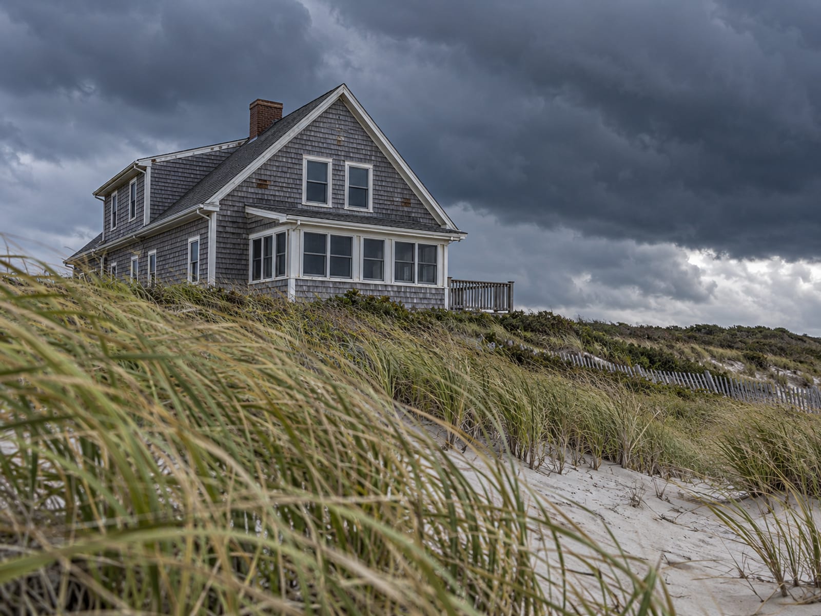 A shingled coastal house under heavy gray clouds with wind-bent beach grass in front
