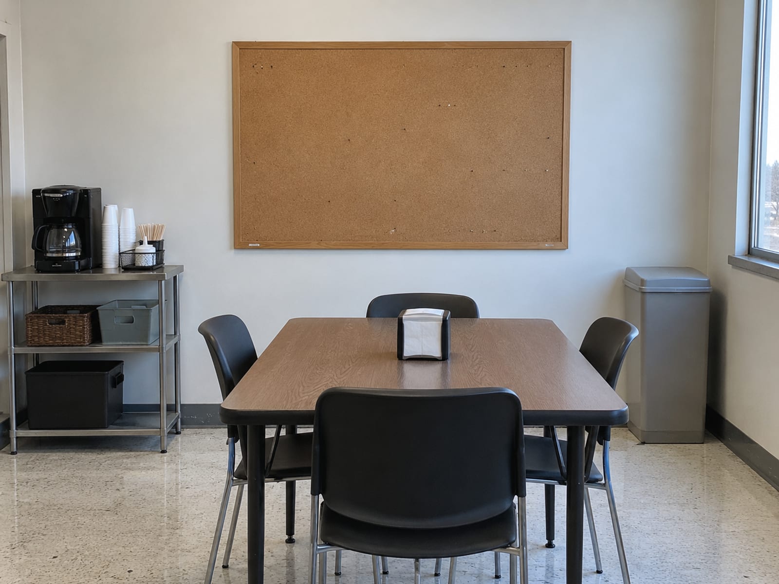 A blank notice board on a break-room wall above a small table with a coffee maker