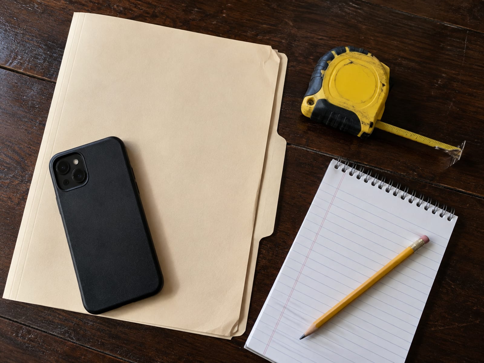 A kitchen table with a closed folder, a phone set to camera mode, a tape measure and a notepad for listing damaged items