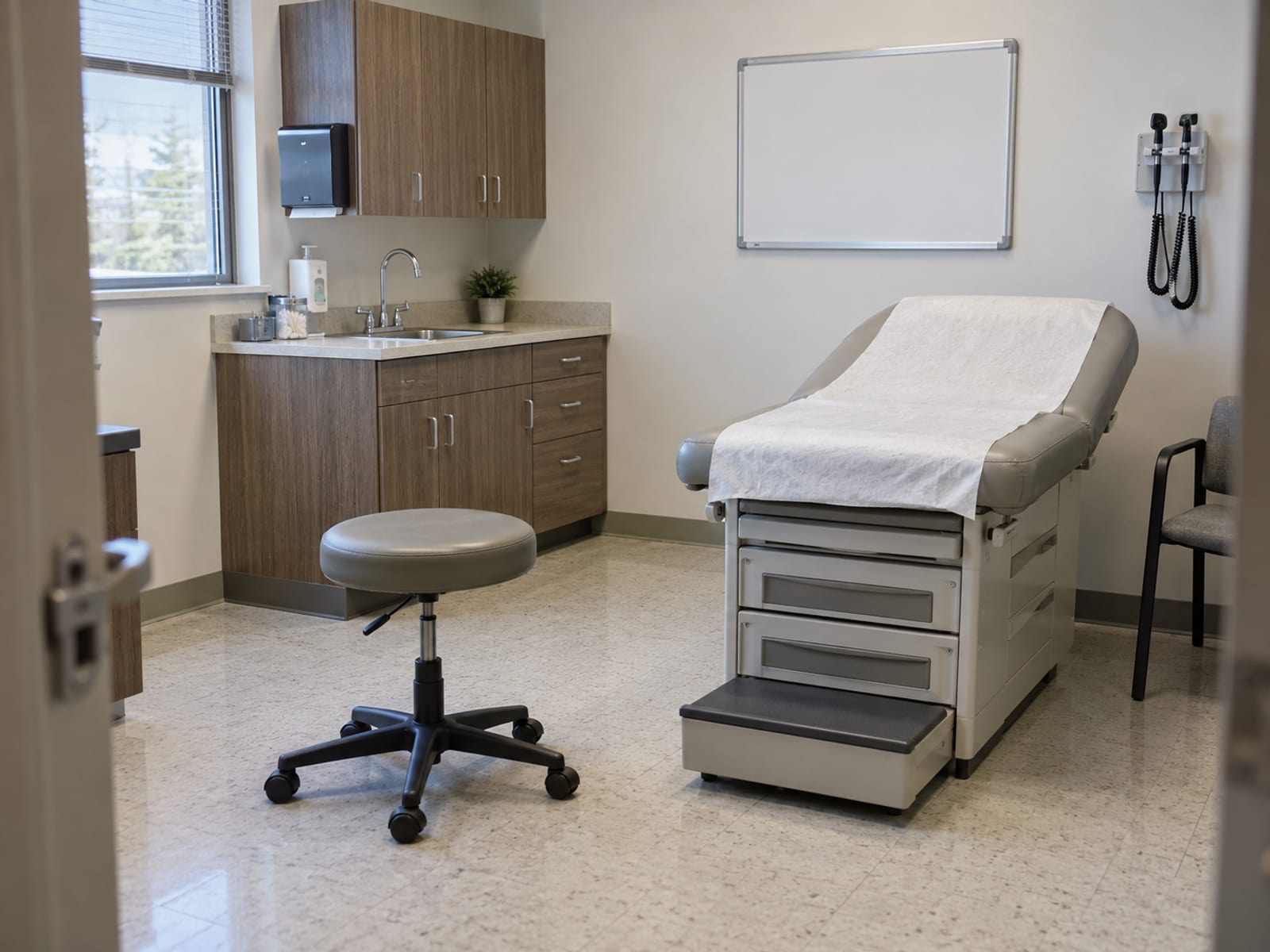An empty clinic examination room with a padded table, a stool and soft window light