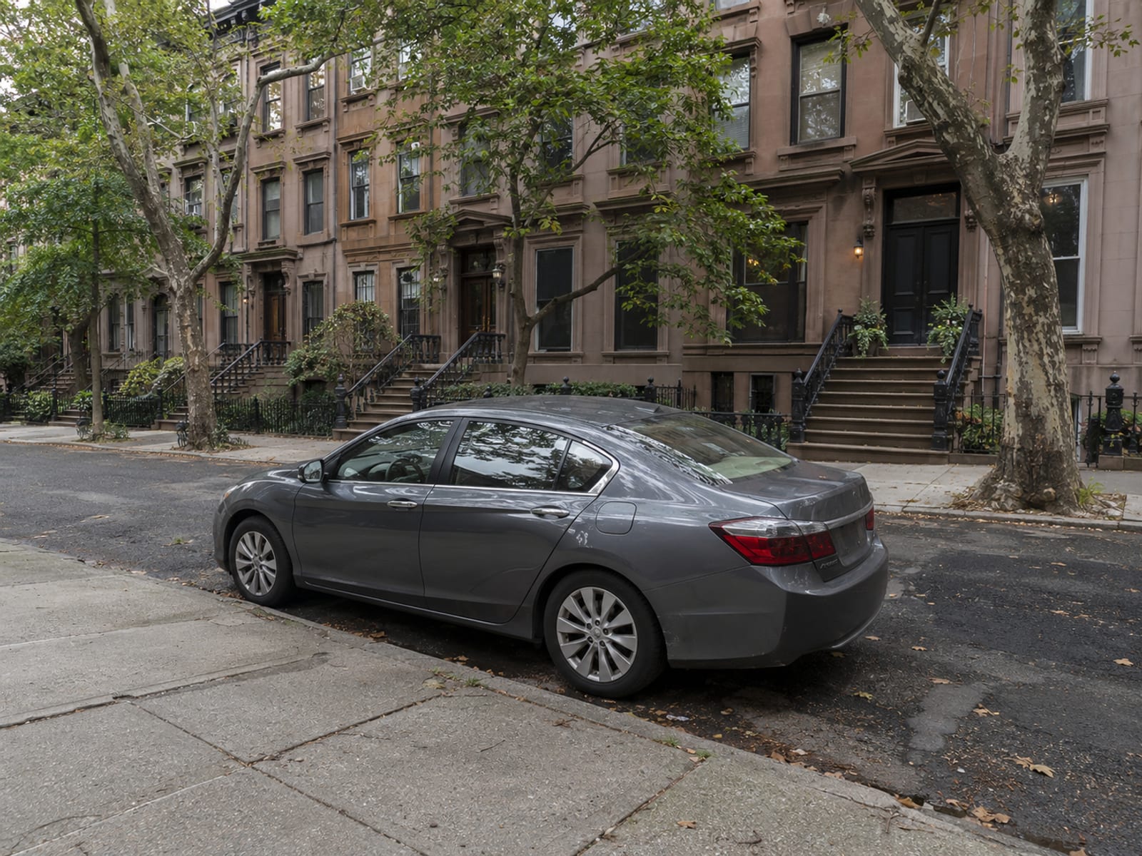 Un sedán estacionado junto a la acera en una calle arbolada de Nueva York con casas de piedra rojiza, visto de lado