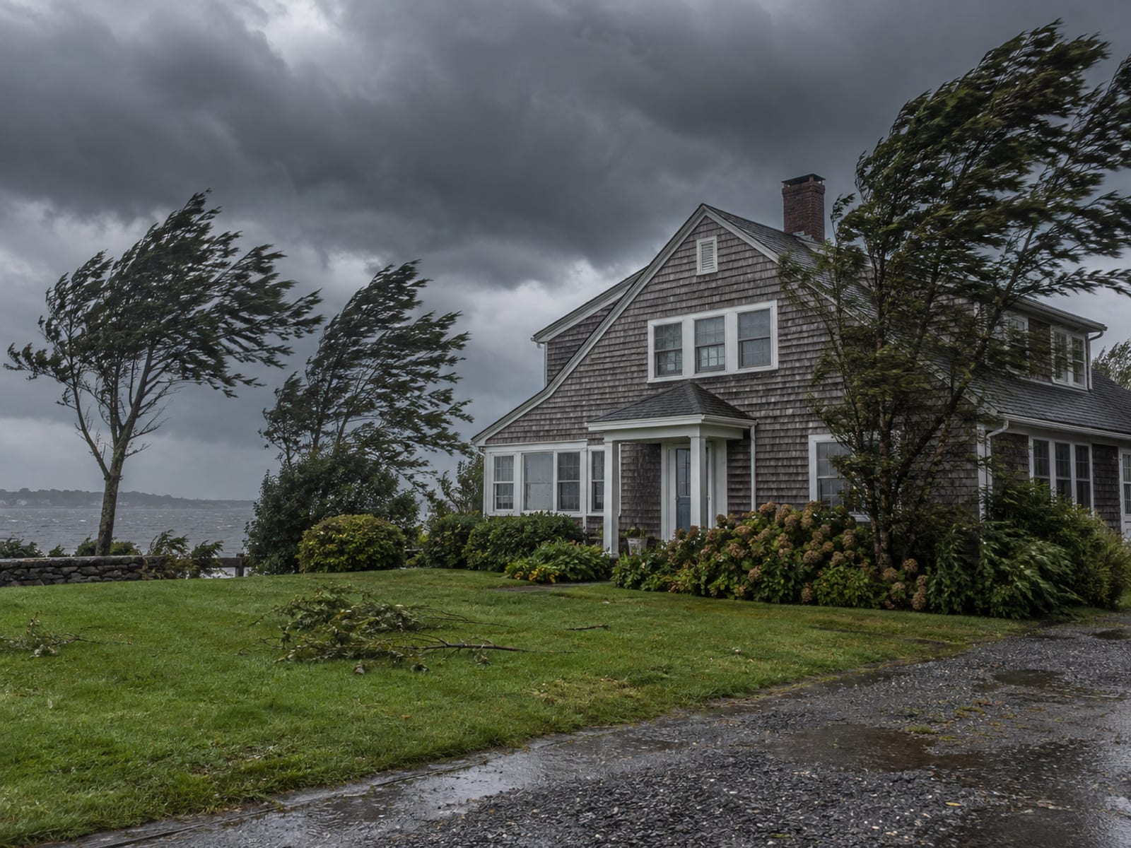 A shingled coastal home under dark storm clouds with bending trees and a wet lawn