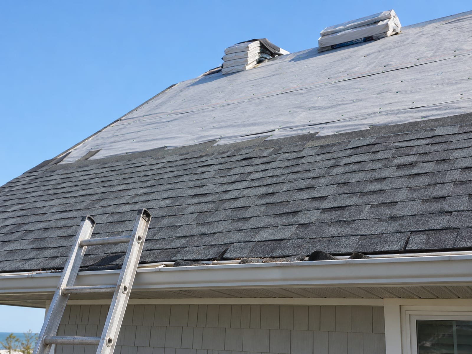 A coastal home roof partway through reshingling, with new shingles, shingle bundles and a ladder under a clear blue sky