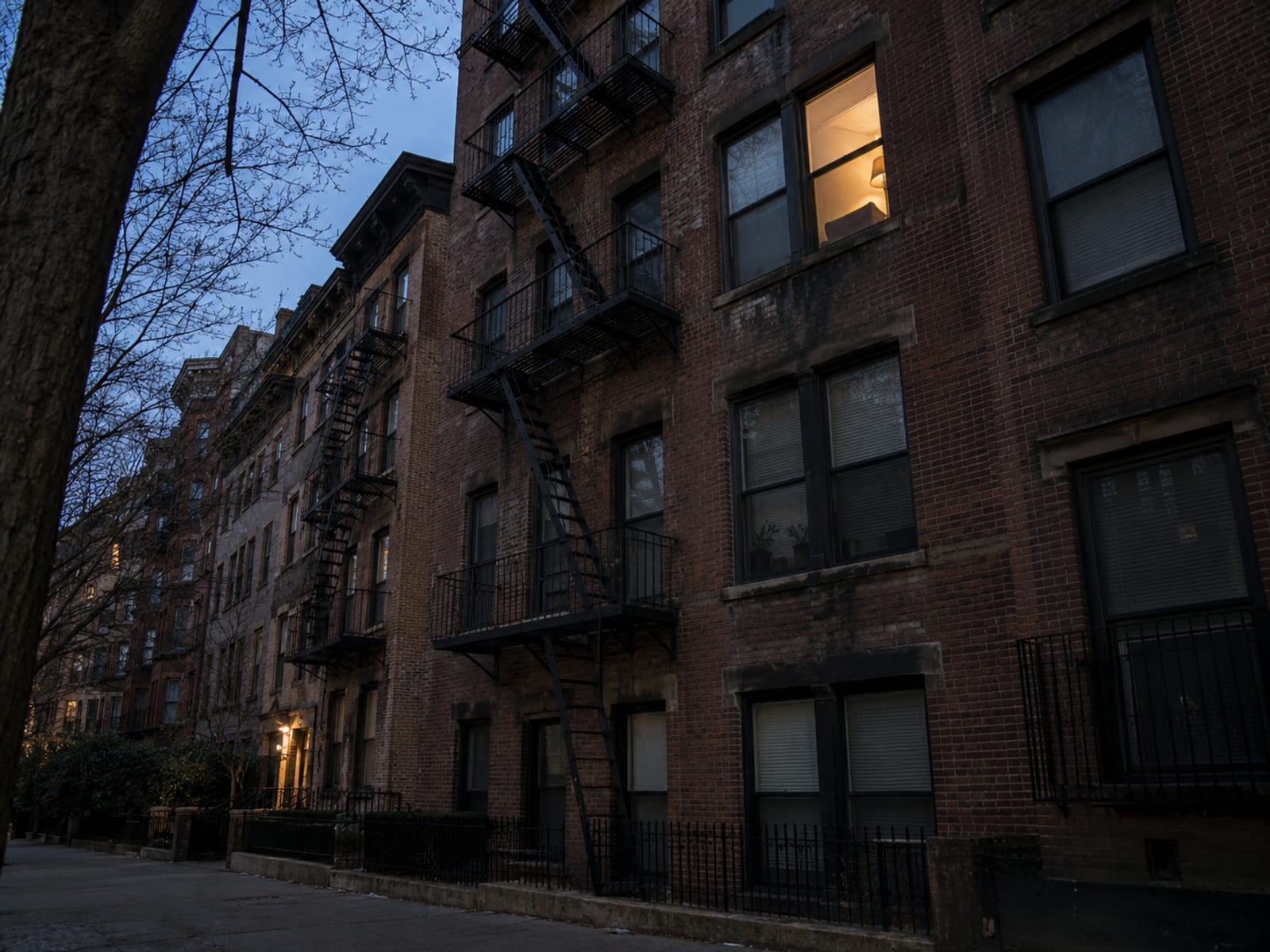 A pre-war brick apartment building seen from the street, with a tenant's sofa and shelves visible through one lit window