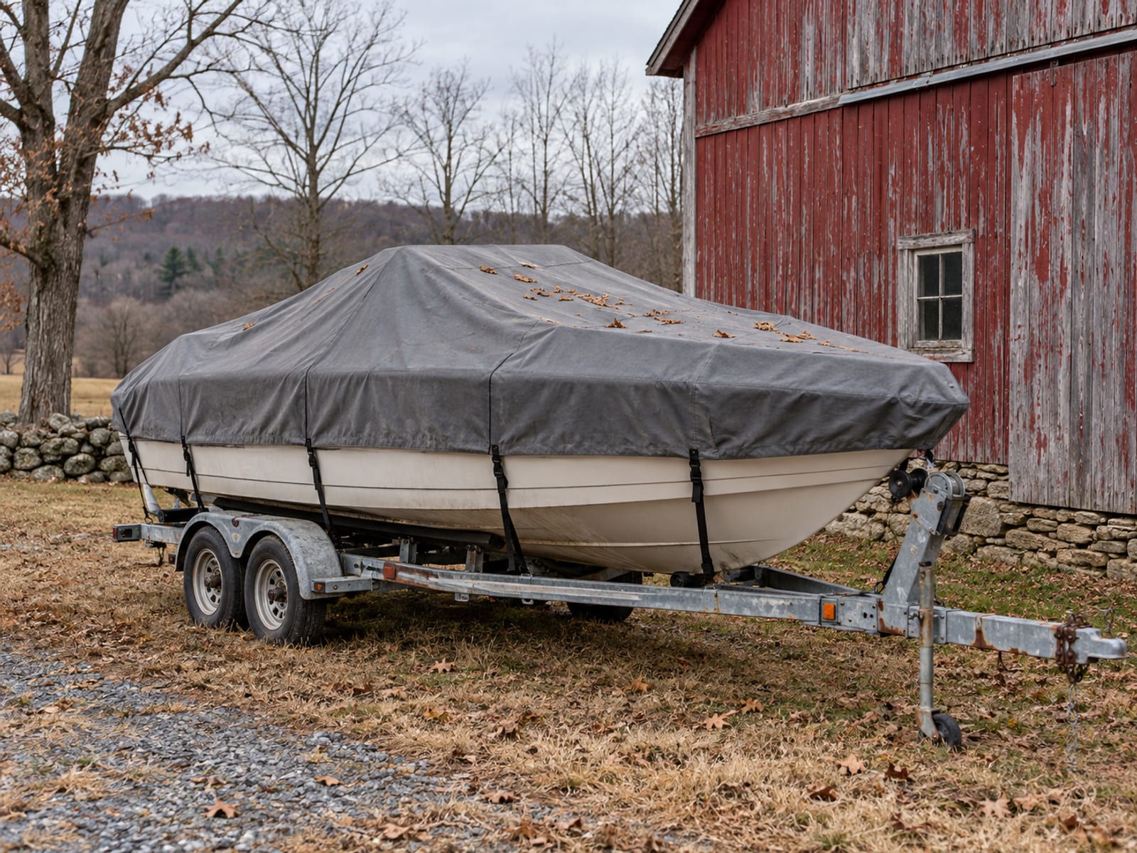 A covered motorboat on a trailer parked beside a barn in late autumn with bare trees around it