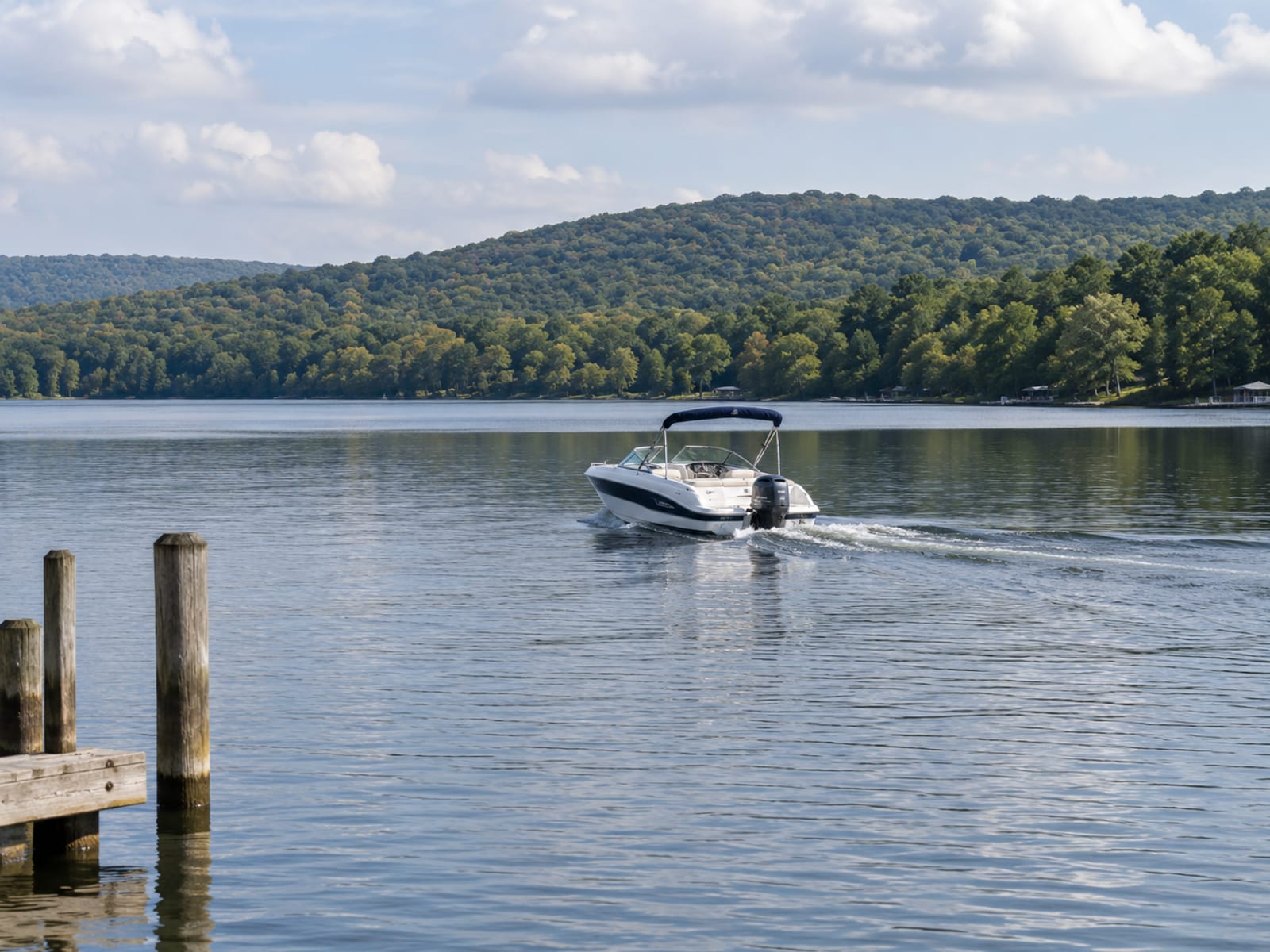 A small motorboat moving slowly past a wooden dock on a calm lake with wooded hills behind it