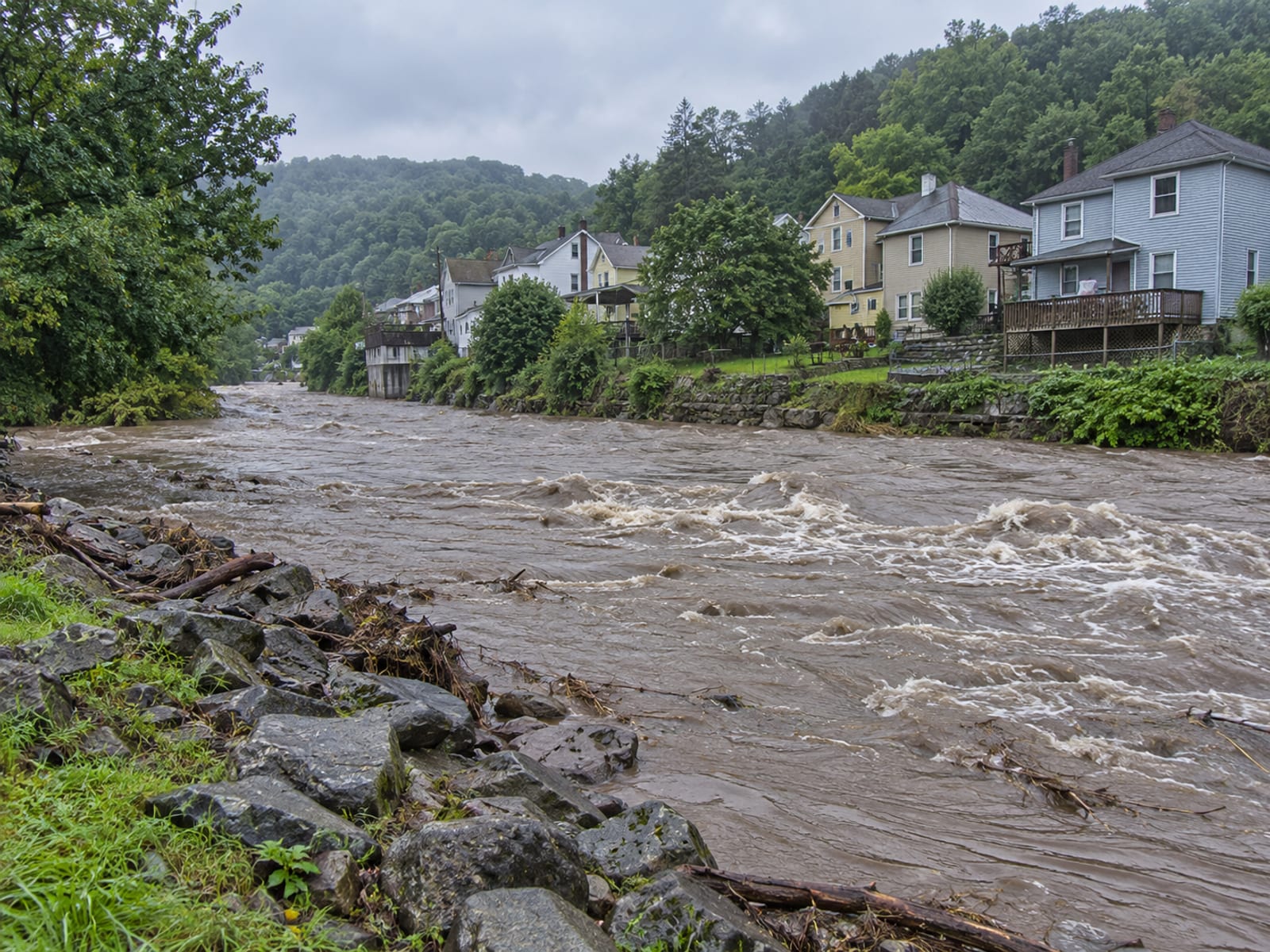 A creek running high beside a row of houses in a small valley town under a gray sky