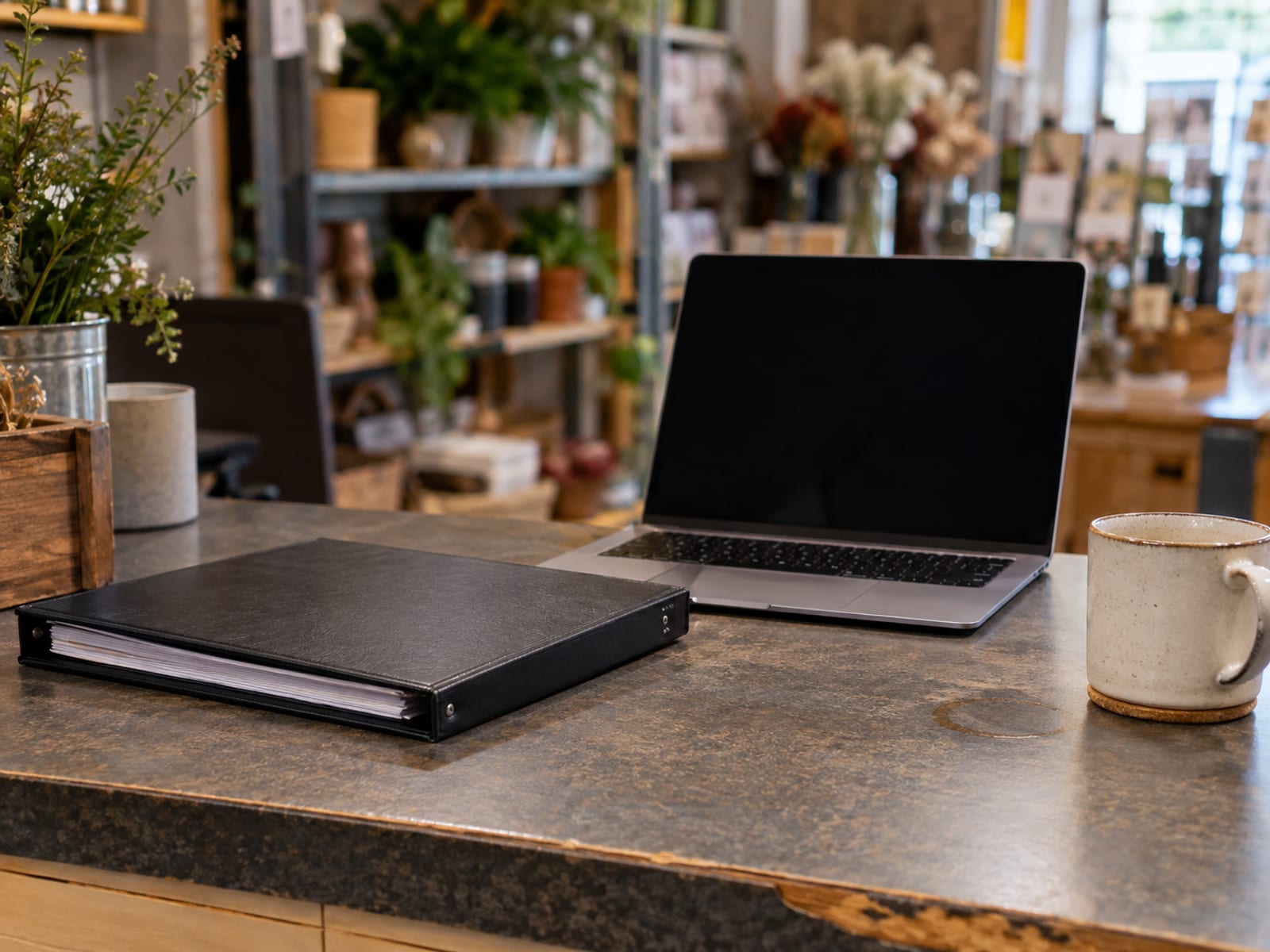 A small shop counter with a closed binder, a laptop and a cup of coffee before opening time