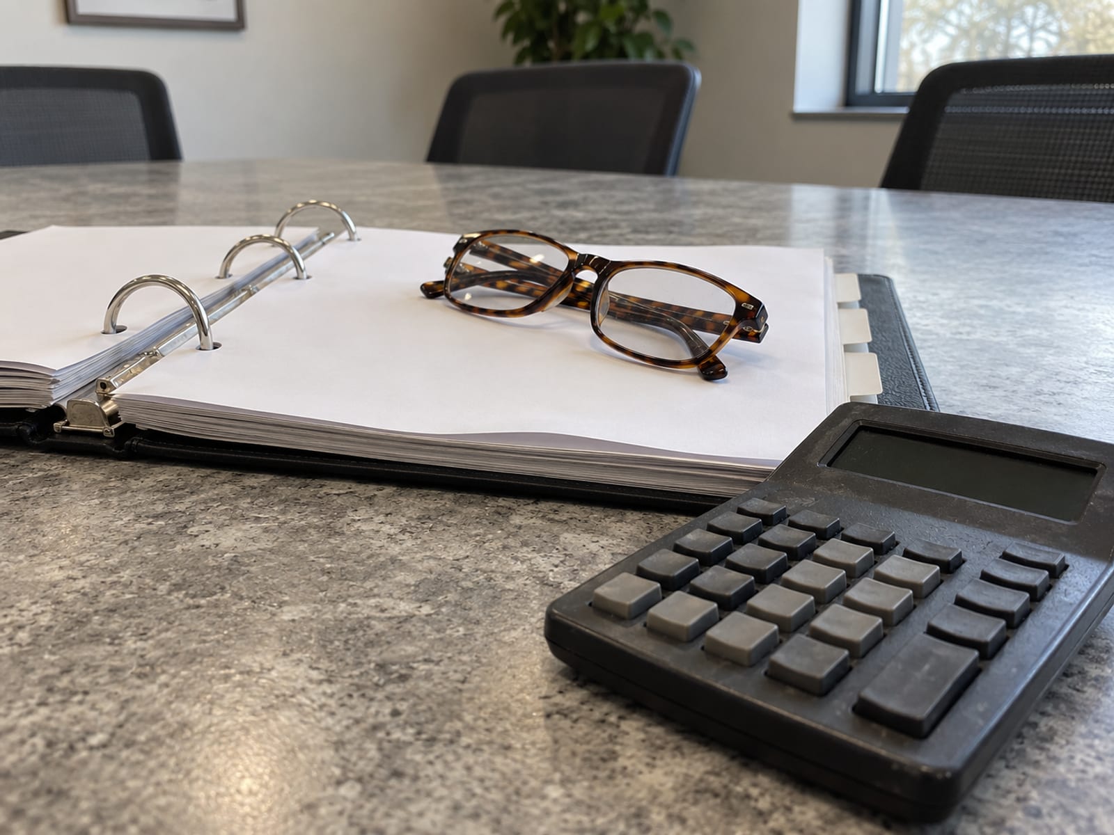 Una carpeta de argollas abierta con páginas en blanco junto a una calculadora y unos lentes de lectura sobre una mesa de juntas