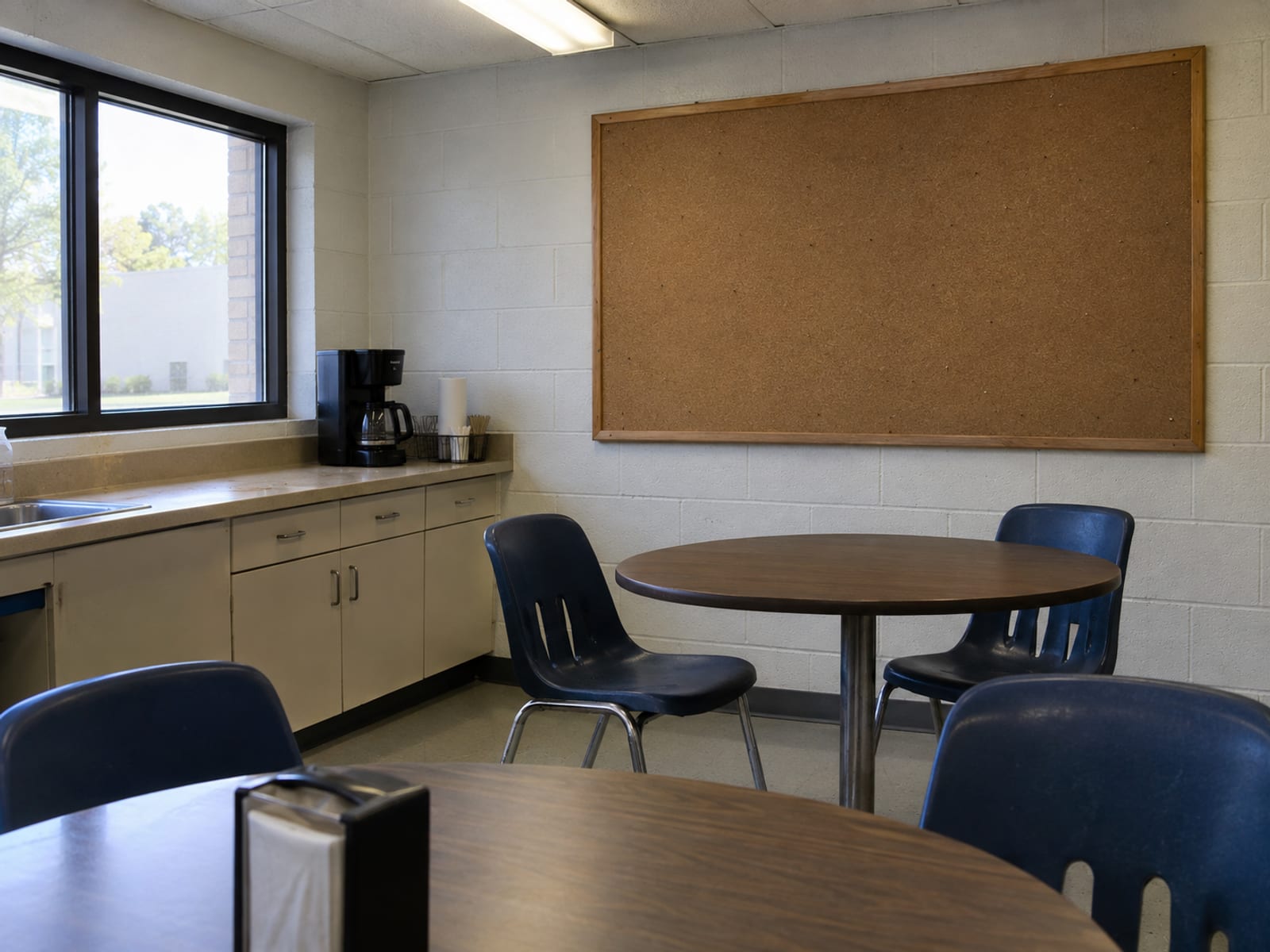 A small office break room wall with a blank notice board, a coffee maker and a few chairs