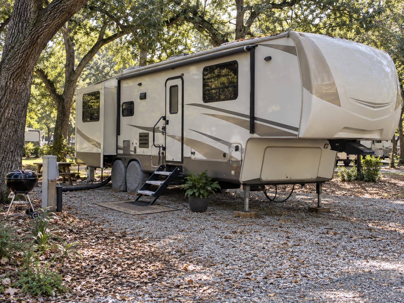 A fifth-wheel trailer parked long term under shade trees on a gravel pad, steps down and no people present