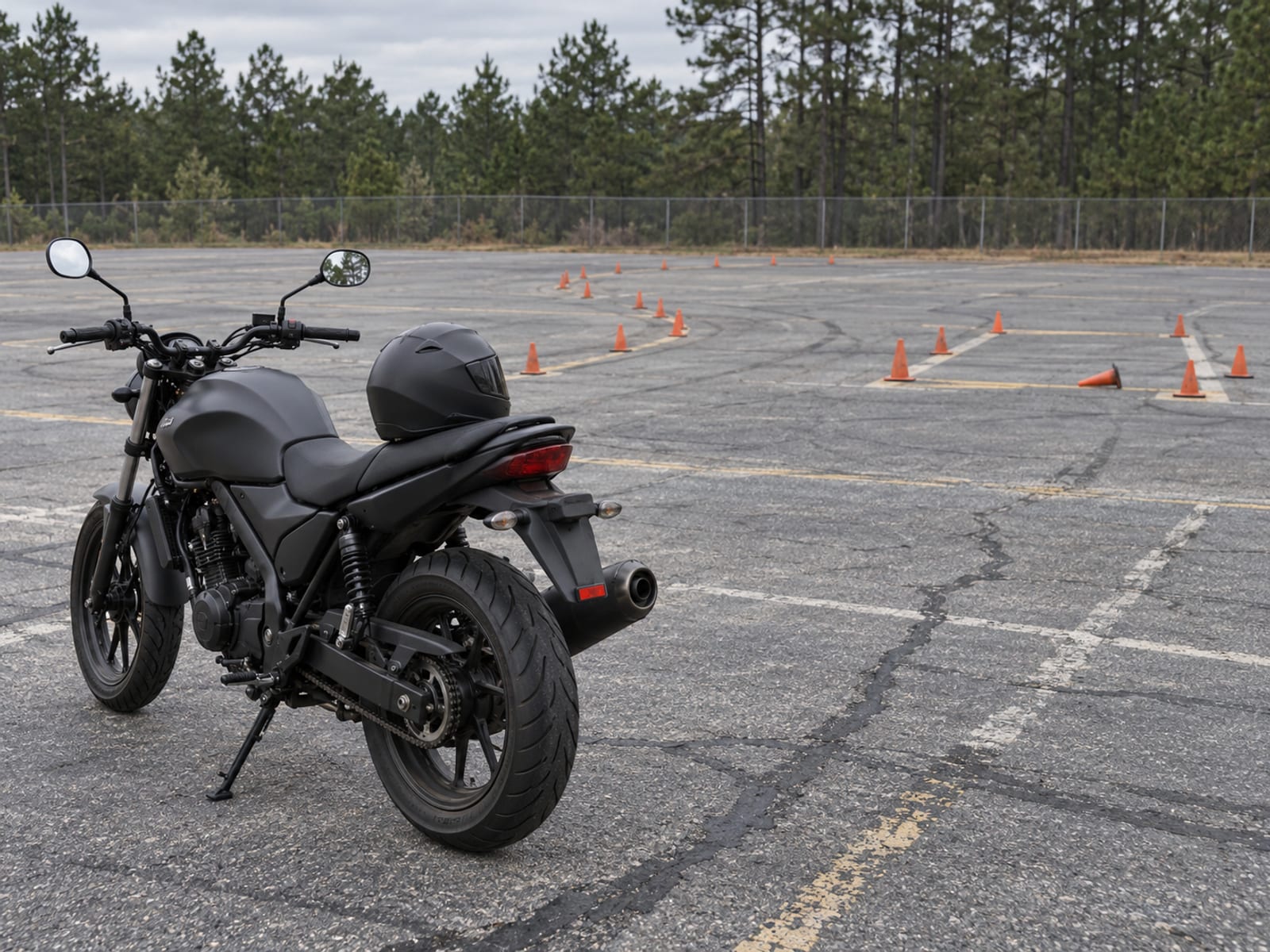 A motorcycle parked on its kickstand beside a weave course of orange cones on an empty paved training lot
