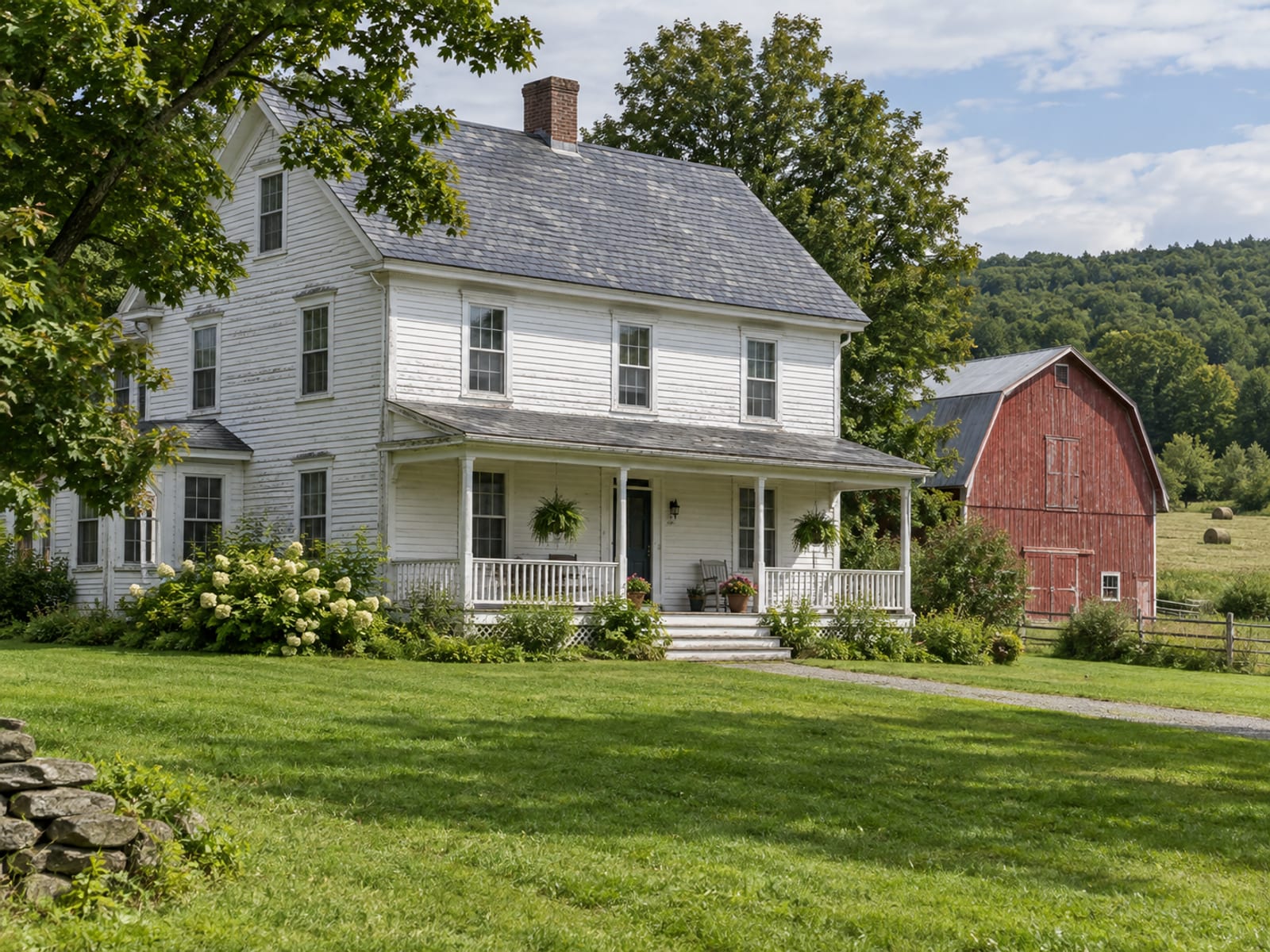 An older white Vermont farmhouse with a slate roof, a porch and a red barn behind it